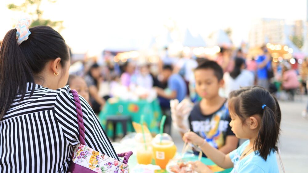 A family enjoying an outdoor event with food and drinks. A woman with a striped shirt and floral bag is seated with two children, a boy and a girl, who are eating at a table. The background is blurred, showing a lively crowd, market stalls, and warm lighting. Mae teulu yn mwynhau digwyddiad awyr agored gyda bwyd a diodydd. Mae menyw mewn crys streipiog a bag blodau yn eistedd gyda dau blentyn, bachgen a merch, sy'n bwyta wrth fwrdd. Mae'r cefndir yn aneglur, yn dangos torf fywiog, stondinau marchnad, a goleuadau cynnes.