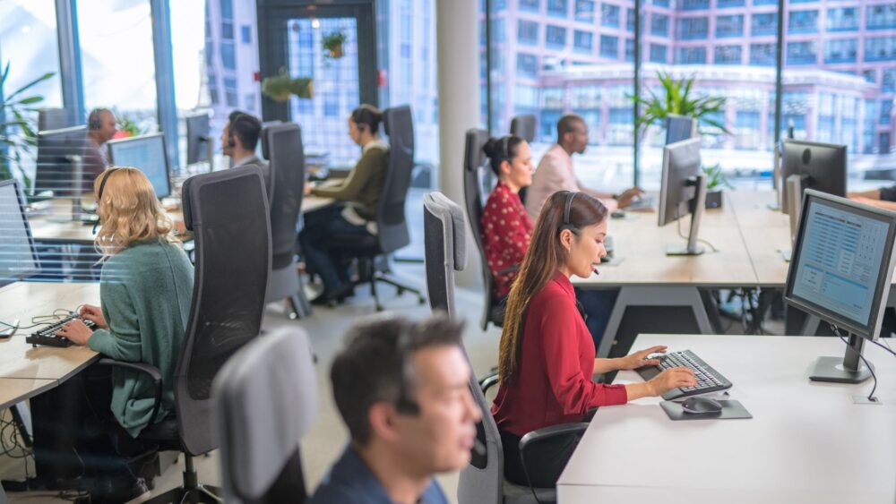 A photo of workers in a call centre sat at their computers working Llun o weithwyr mewn canolfan alwadau yn eistedd wrth eu cyfrifiaduron yn gweithio