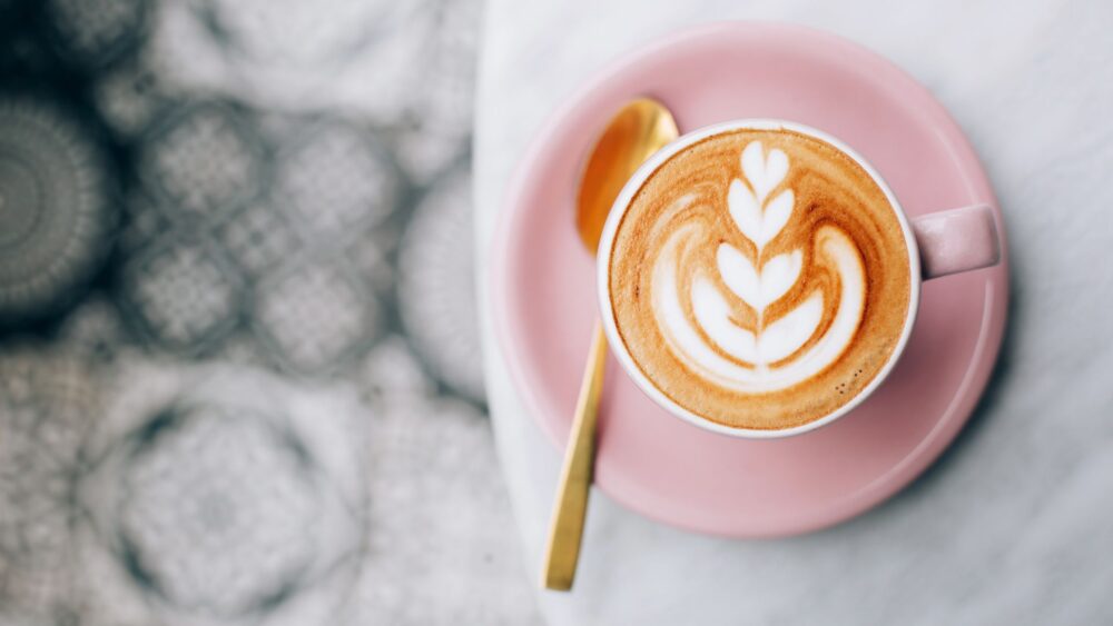 A pink cup of coffee with intricate latte art resembling a leaf design, served on a matching pink saucer with a golden spoon, set against a blurred decorative patterned background.