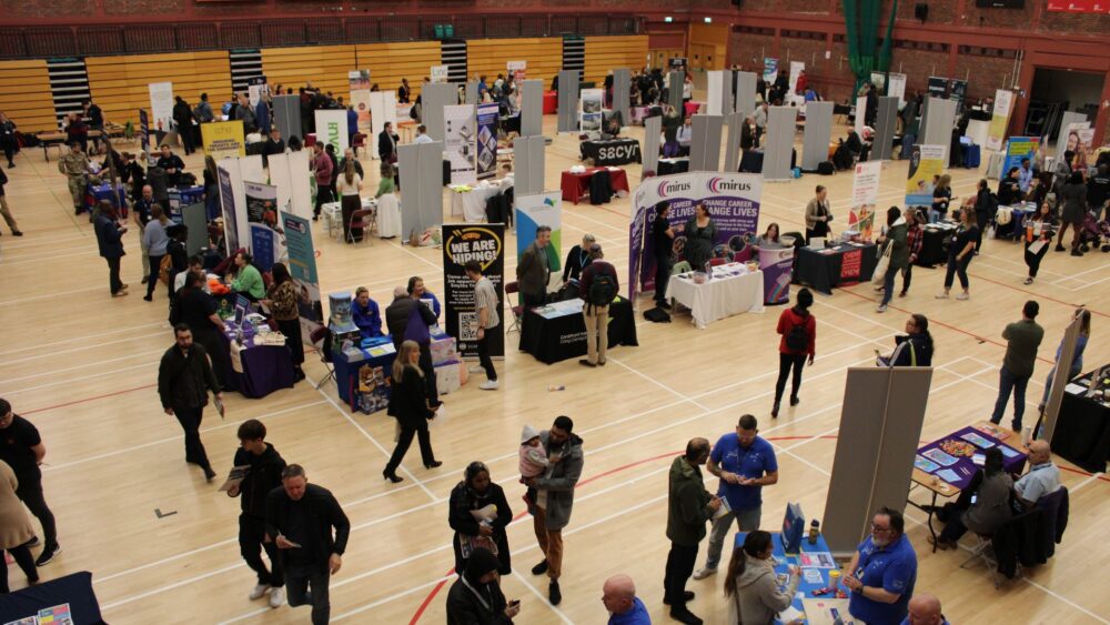 A bustling job fair held in a large indoor gym with rows of booths and displays set up by various organisations. Attendees, including job seekers and representatives, interact and network around the booths. The floor is marked with sports court lines, and the space is filled with activity, as people explore career opportunities.