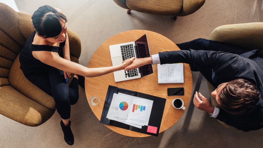 An image of two people shaking hands over a small coffee table Delwedd o ddau berson yn ysgwyd llaw dros fwrdd coffi bach