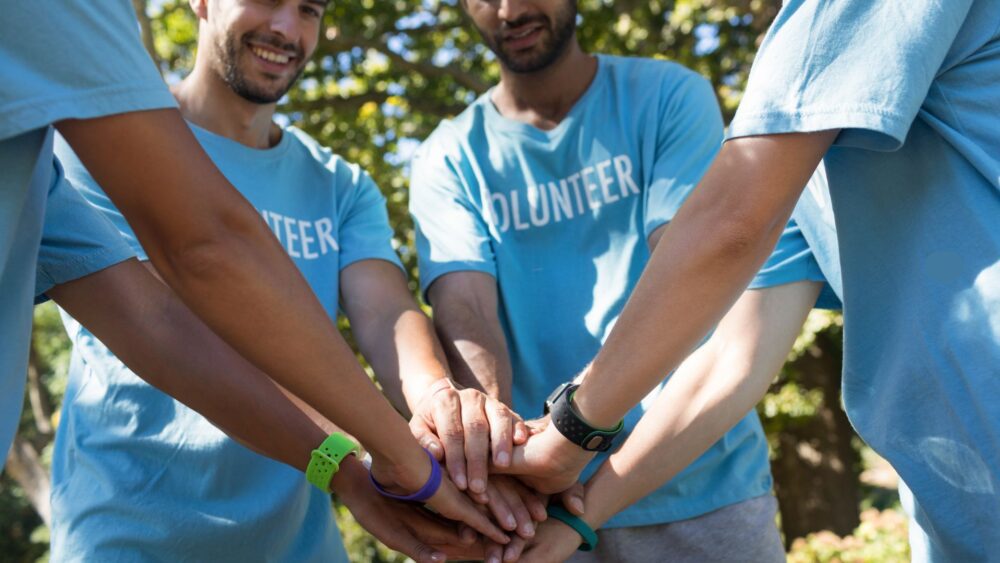 A group of volunteers wearing matching light blue t-shirts with the word "VOLUNTEER" printed on them stand in a circle outdoors. They place their hands together in a unified stack, symbolizing teamwork and cooperation. The background consists of lush green trees, indicating an outdoor setting with natural sunlight. Mae grŵp o wirfoddolwyr yn gwisgo crysau-T glas golau cyfatebol gyda’r gair "VOLUNTEER" wedi’i argraffu arnynt yn sefyll mewn cylch yn yr awyr agored. Maent yn gosod eu dwylo gyda’i gilydd mewn pentwr unedig, yn symbol o waith tîm a chydweithrediad. Mae'r cefndir yn cynnwys coed gwyrdd toreithiog, gan nodi lleoliad awyr agored gyda golau haul naturiol.